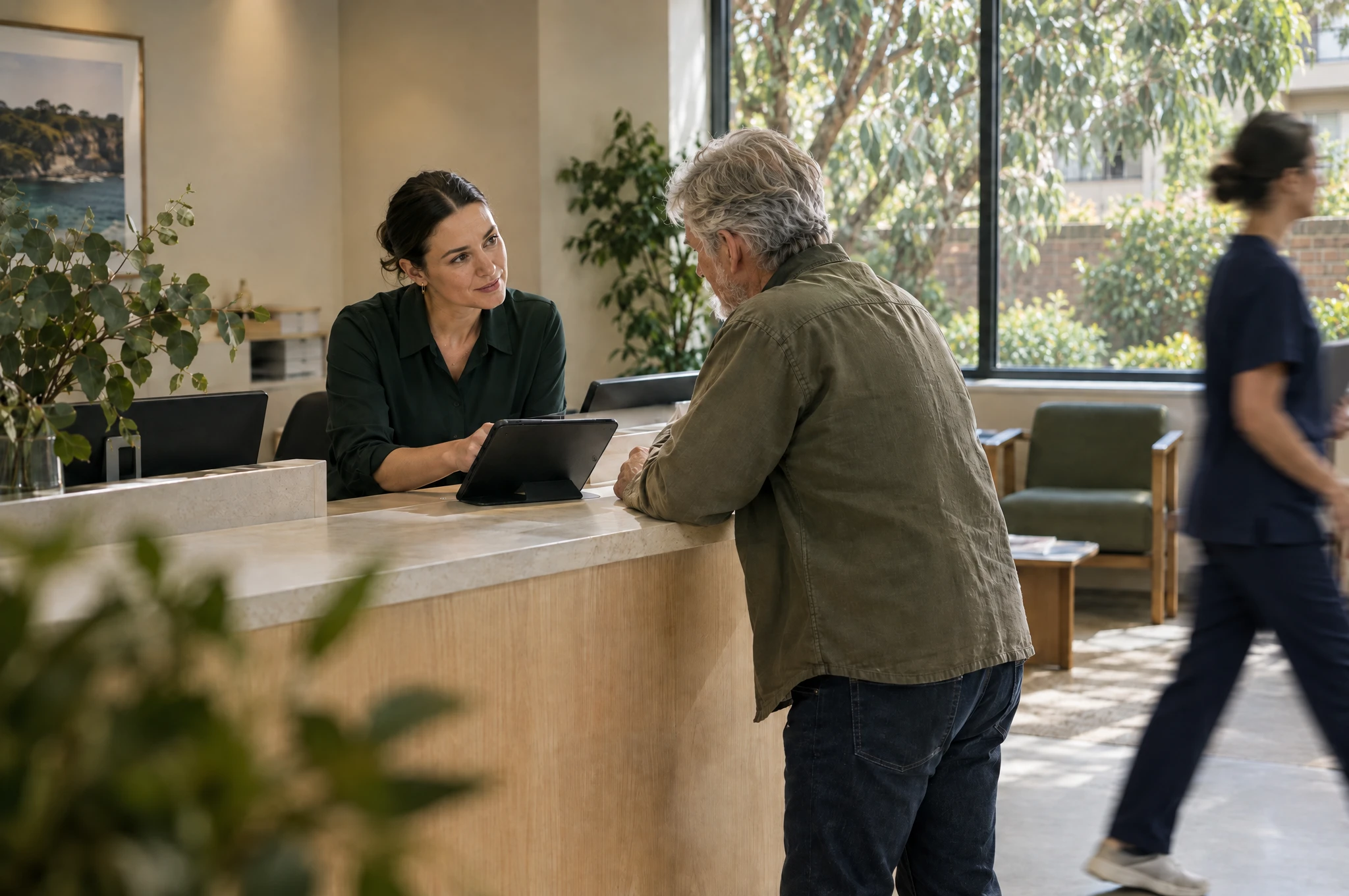 A receptionist explains a tablet to a patient at a clinic’s timber reception desk.